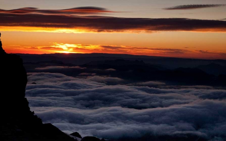 High above clouds, the sun rises or sets, causing the sky to appear in shades of orange and yellow.